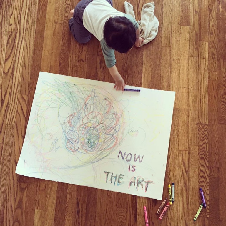 Baby with crayons on a large piece of paper with drawings and the words, NOW IS THE ART