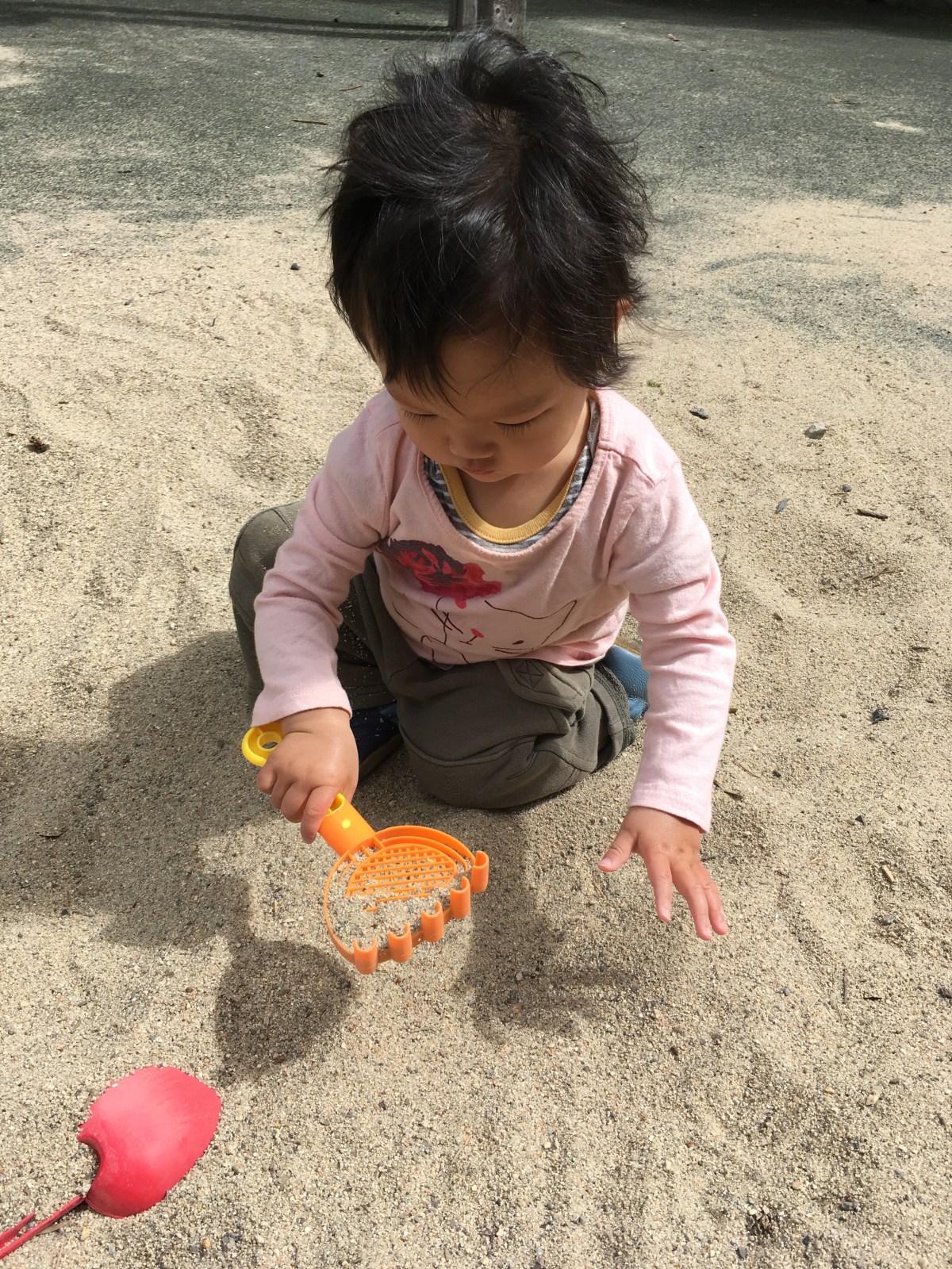 15-month-old Ada, wearing a pink shirt, plays in the sand with an orange rake