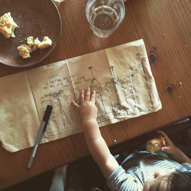 A fineliner drawing on a brown paper napkin, on a table in front of a toddler, whose hand is on it