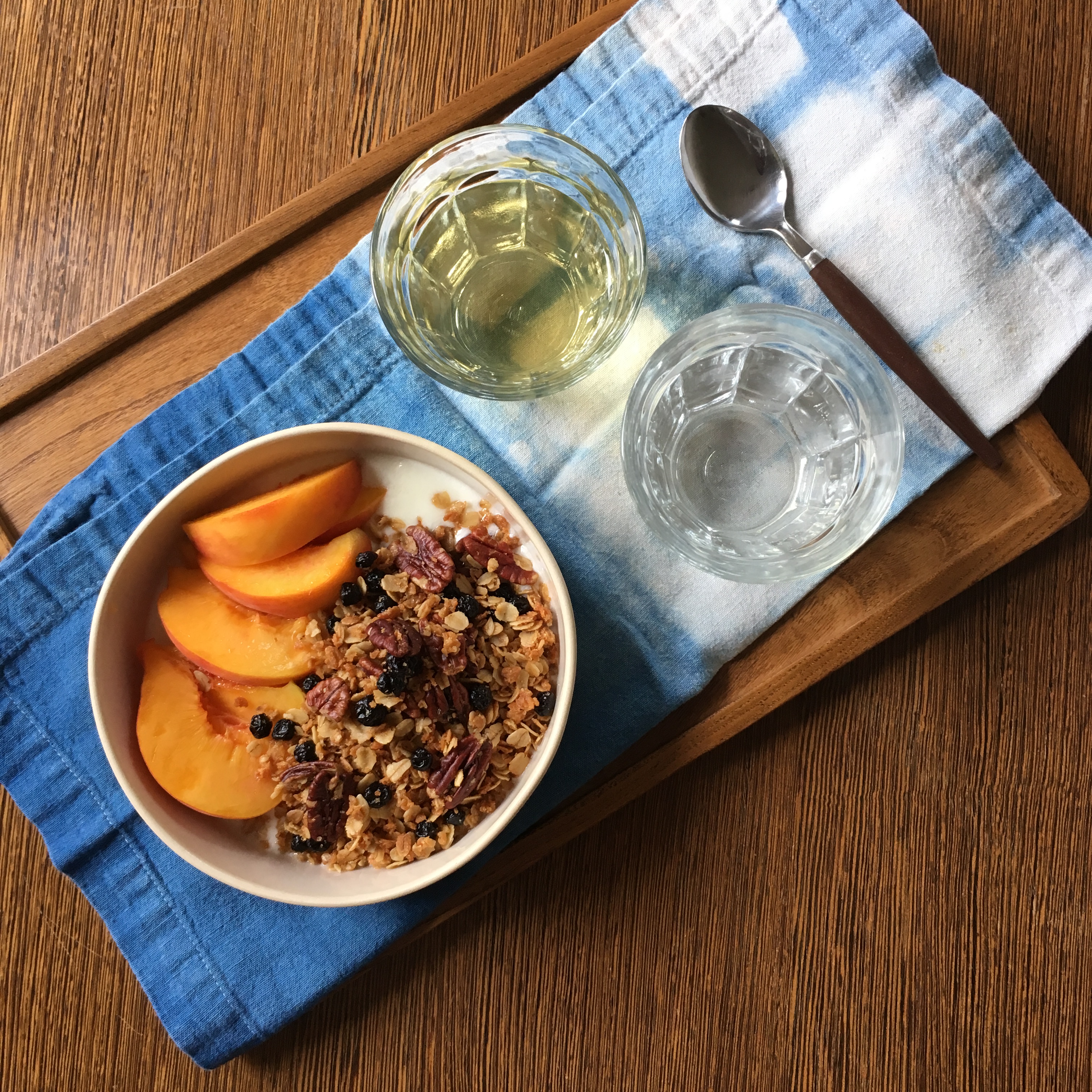 A bowl of peaches, yogurt, and granola, with a spoon, a blue and white napkin, and glasses of water and green tea alongside.