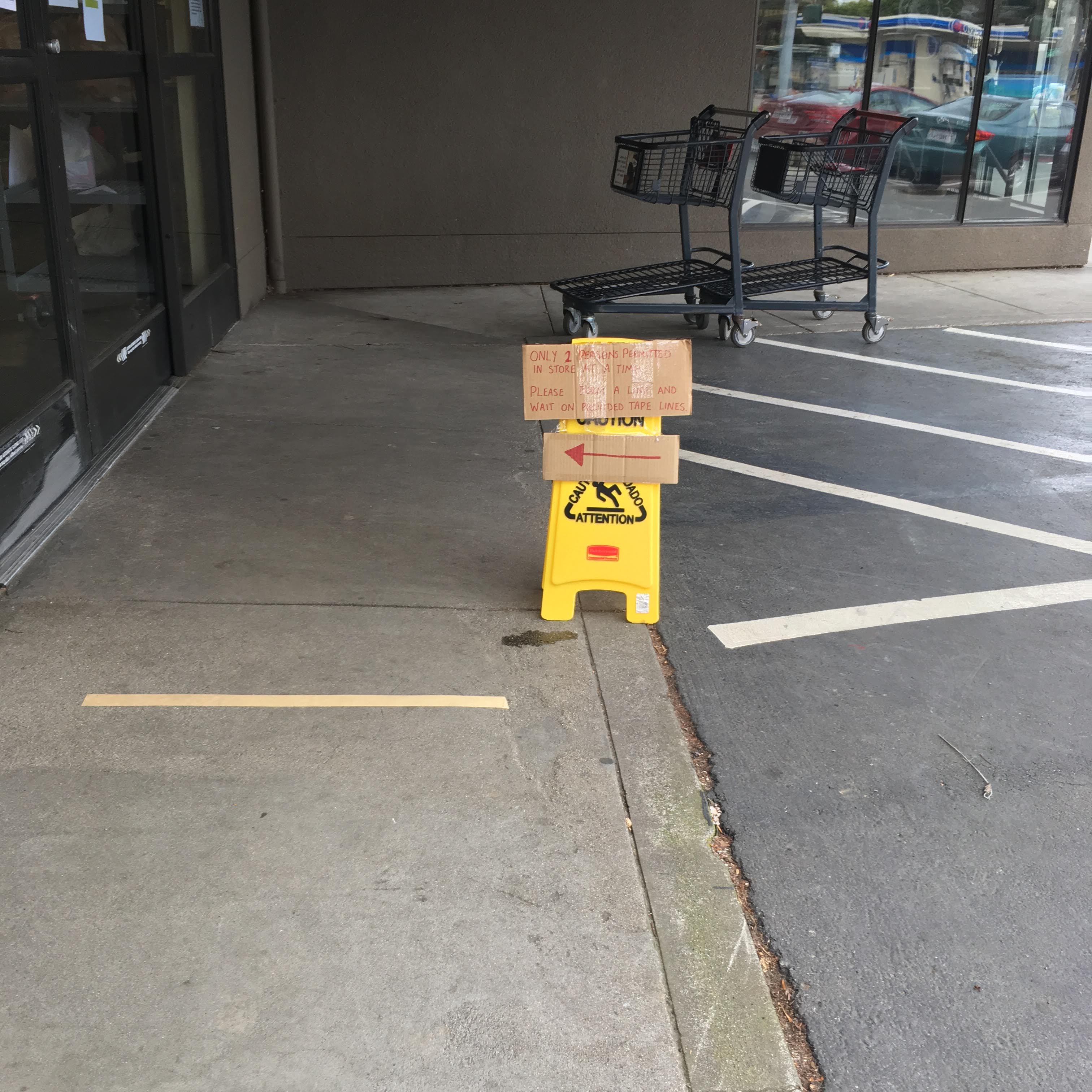 Social-distancing sign in red marker on cardboard, taped to a yellow CAUTION sign