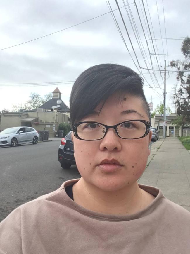 Asian woman with short hair and shaved sides, black glasses, taupe sweatshirt