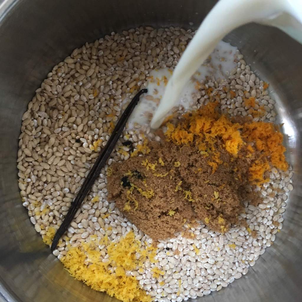 Milk being poured into a pot of pearl barley, brown sugar, citrus zests, and vanilla bean
