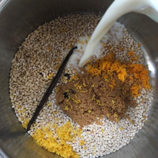 Milk being poured into a pot of pearl barley, brown sugar, citrus zests, and vanilla bean
