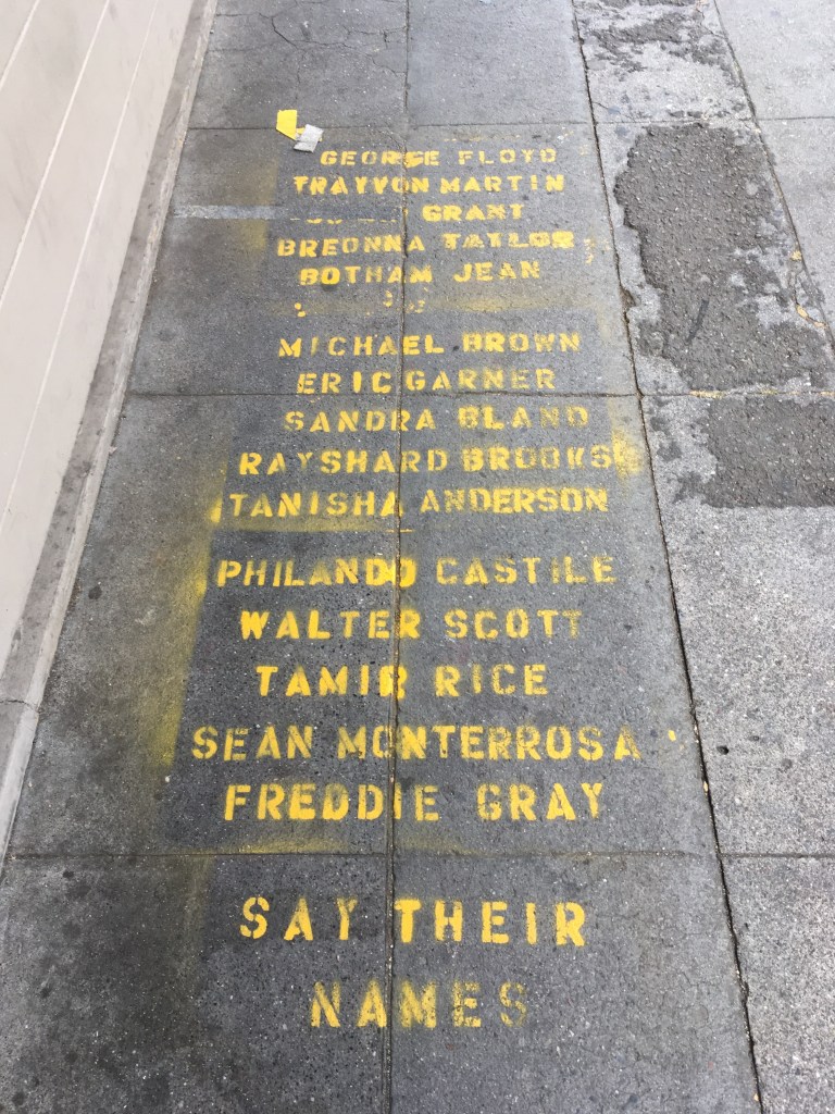 A list of names of Black people killed by police, stencilled in yellow on a sidewalk
