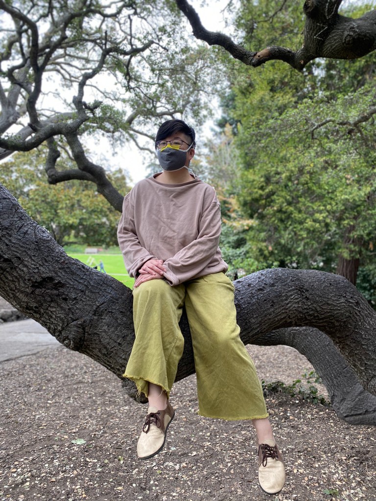 Short-haired Asian American person in face mask, taupe sweatshirt and lace-up shoes, and light green wide-legged pants sits in a tree in Piedmont Park