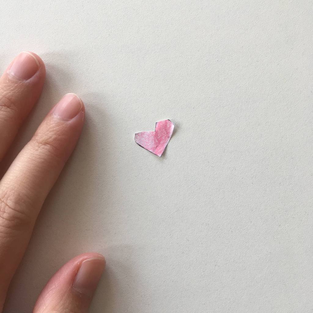 Light-skinned adult hand next to a tiny paper heart crayoned pink