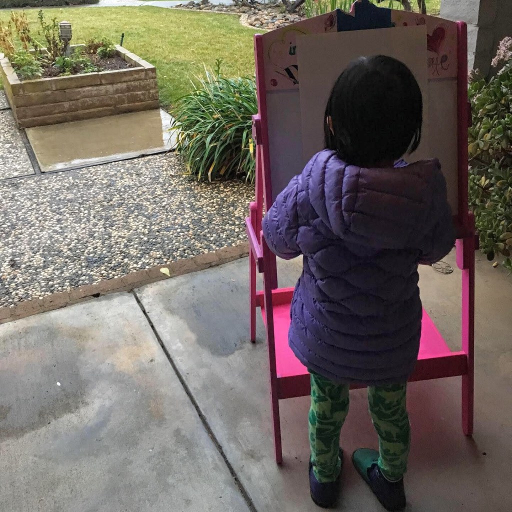 A small dark-haired child draws outdoors at an easel