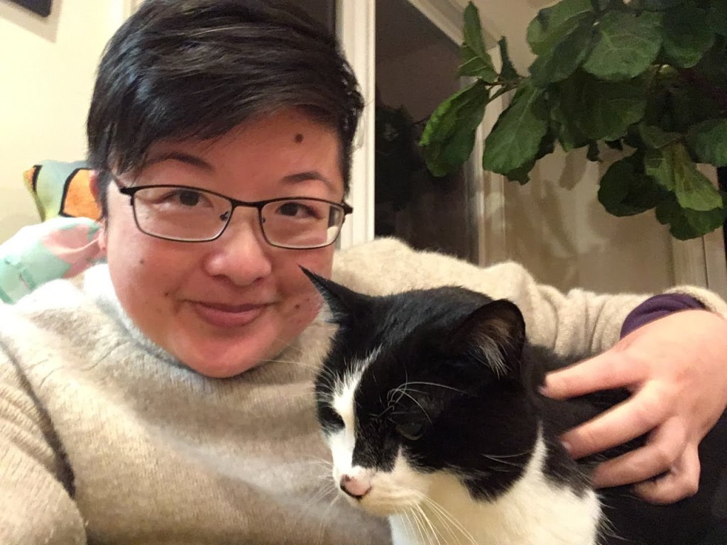 A short-haired Asian person with glasses pets a black and white cat
