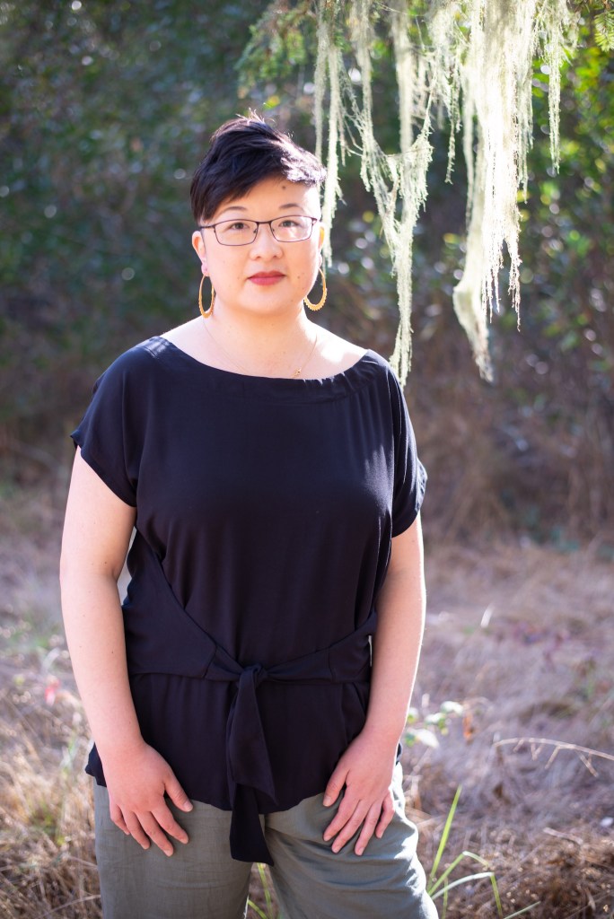 Lisa Hsia, a short-haired Asian person in glasses, stands under a tree in the sunshine